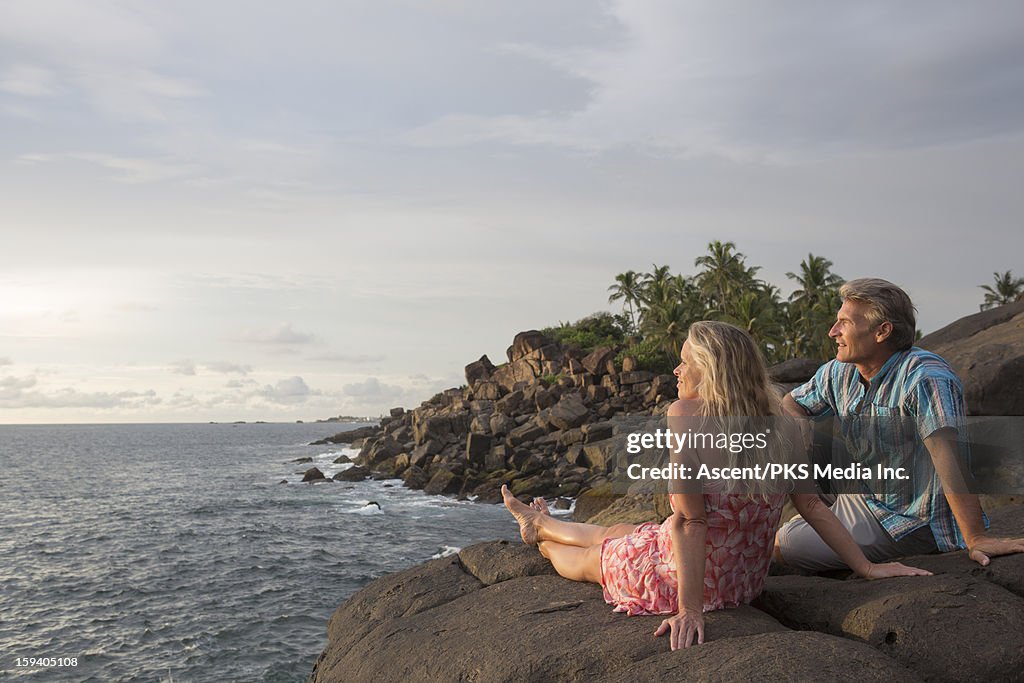Couple sit on rocks above sea, watch sunrise