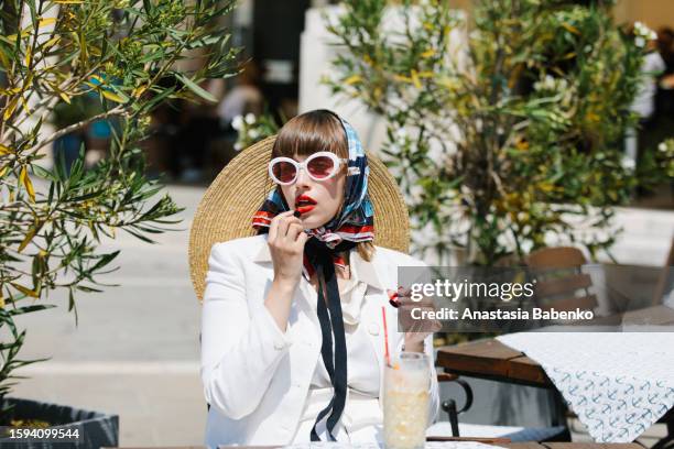 young woman in white suit sitting at sidewalk cafe and putting on red lipstick - woman headscarf stock pictures, royalty-free photos & images