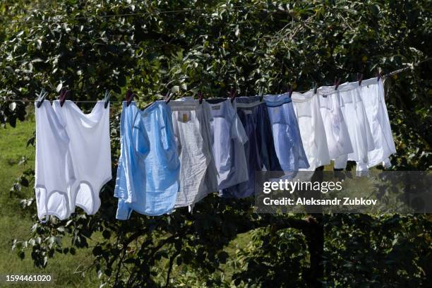 clean sweaters, trousers and t-shirts hang on a rope outside on a sunny summer day, against a blue sky. washed women's, men's and children's clothes, bed linen are dried on a clothesline. the concept of housework, household duties. - women in wet tee shirts stock pictures, royalty-free photos & images