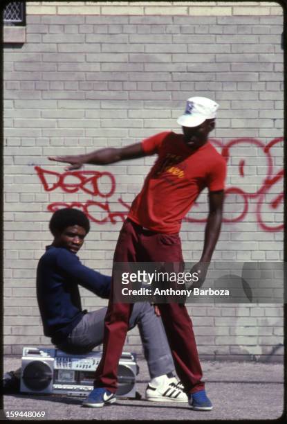 Young man breakdances on a city street while his friend sits on a boombox behind him, 1980s.