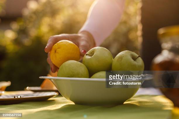 woman putting a fruit bowl with apples and lemons on a table in a sunny back yard - fruitschaal stockfoto's en -beelden