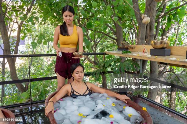 a woman with her eyes closed inside a tub of ice doing cryotherapy while her coach plays a singing bowl - bad breath stockfoto's en -beelden
