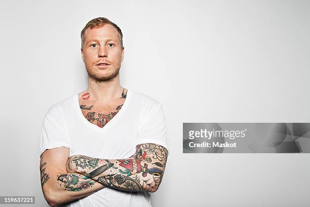 portrait of a tattooed man with arms crossed standing against grey background - tattoo stock-fotos und bilder