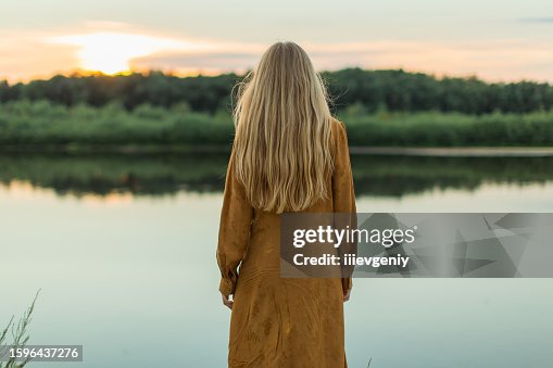 Rückansicht. Blonde Frau im gelben Kleid, die sich am Flussufer ausruht. Sommertag. Lange Haare. Sand. Rückansicht. Blonde Frau im gelben Kleid, die sich am Flussufer ausruht. Sommertag. Lange Haare. Sand.