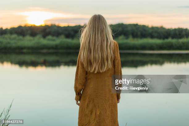rear view. blonde woman in yellow dress resting on the river bank. summer day. long hair. sand. - lang haar stockfoto's en -beelden