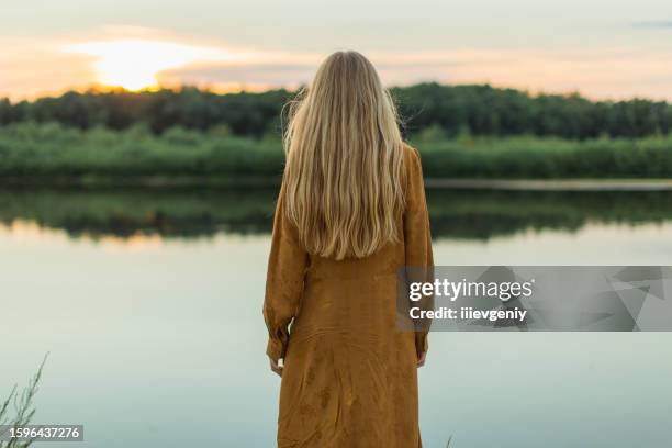 rear view. blonde woman in yellow dress resting on the river bank. summer day. long hair. sand. - long river stock pictures, royalty-free photos & images