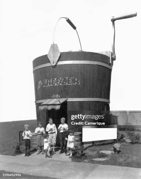 Handful of children standing out front of an ice cream store named The Freezer in the shape of an old fashion bucket with a crank.