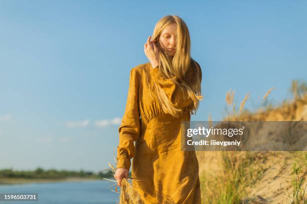 blonde woman in yellow dress resting on the river bank. sunset light. summer day. long hair. sand. broken heart - woman long dress beach stock pictures, royalty-free photos & images