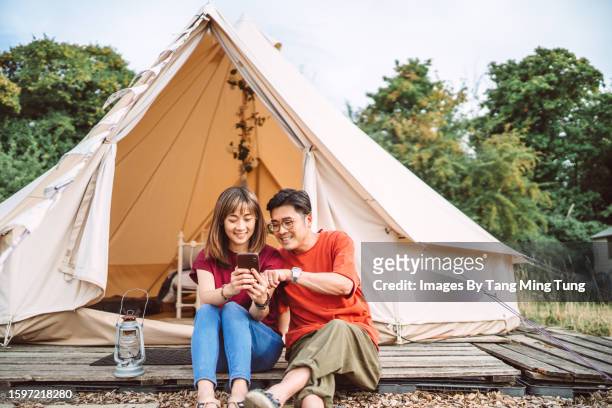 young couple using smart phone together joyfully while sitting on the wood deck outside a glamping dome tent - zoom meeting stock pictures, royalty-free photos & images