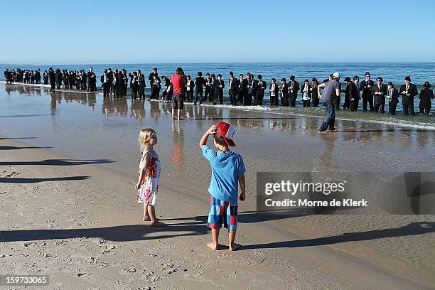 Spectators look on while people stand on the beach in suits and bowler hats as part of an art installation created by surrealist artist Andrew Baines...