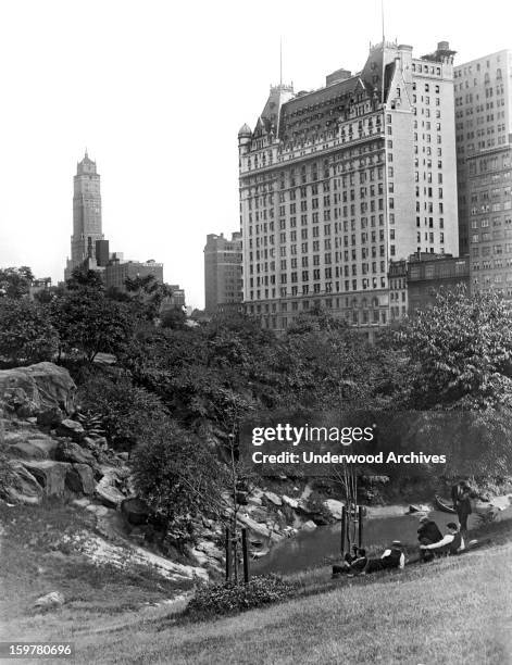 The famed Plaza Hotel rises from the trees in Manhattan's lovely Central Park, New York, New York, c. 1928. Five young men relax along the banks of...