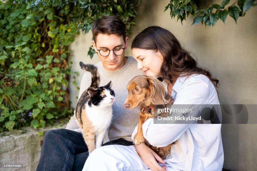Young couple sitting in backyard holding a cat and a dog