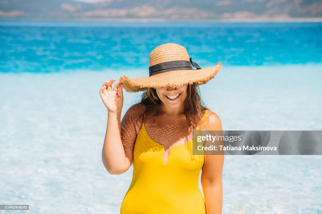 A woman in a yellow swimsuit and straw hat emerges from the sea with clear turquoise water on a summer sunny day.