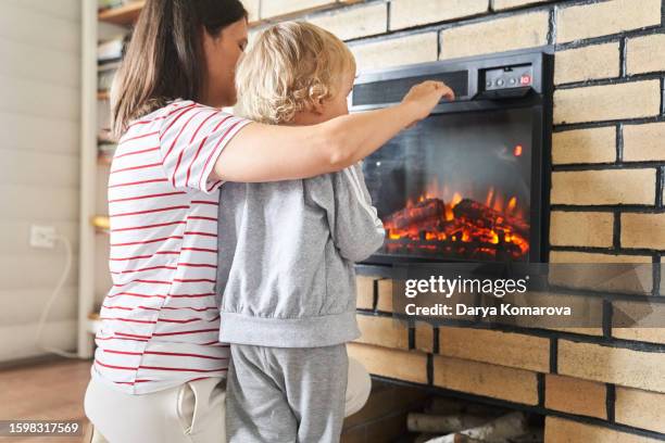 mom and son spend time in the house by the fireplace, electric fireplace as a heater, happy people, lifestyle photos with copy space. - fireplace stock pictures, royalty-free photos & images