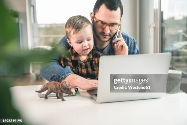 father working at table in home office with son sitting on his lap - kompatibilität stock-fotos und bilder