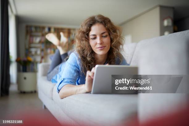 beautiful woman using digital tablet while lying on sofa at home - auf dem bauch liegen stock-fotos und bilder