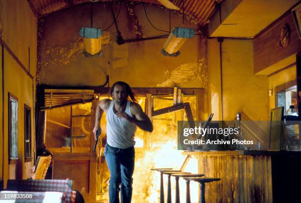 Nicolas Cage running through a burning building while holding onto a gun in a scene from the film 'Con Air', 1997.