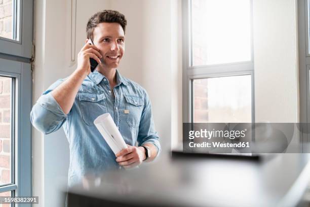 smiling businessman on cell phone at the window in office - rolling up sleeve stock-fotos und bilder
