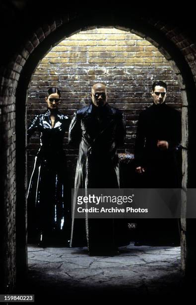 Carrie-Anne, Moss Laurence Fishburne, and Keanu Reeves standing against brick wall in a scene from the film 'The Matrix Reloaded', 2003.