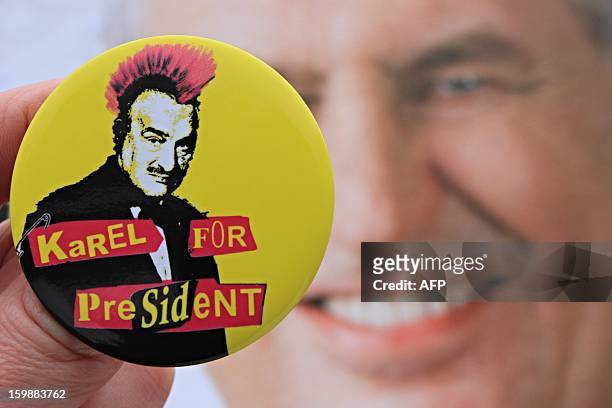 Hand holds a badge depicting Czech Presidential candidate Karel Schwarzenberg as a punk with red hair in front of a campaign poster of his contender...