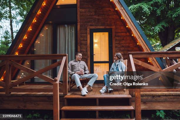 glamping retreat and fresh woodland air for romantic atmosphere. man and woman enjoy conversation near illuminated log cabin outdoors - casita de campo fotografías e imágenes de stock