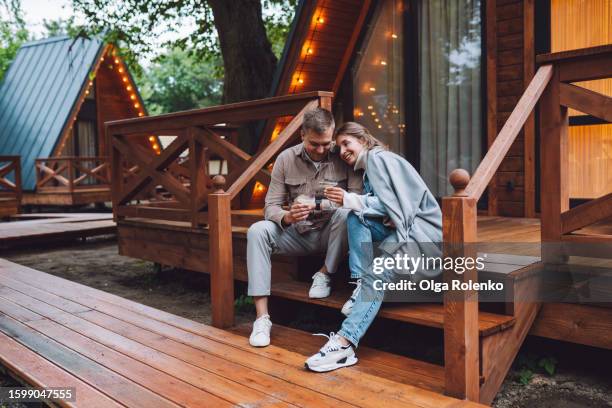 romantic getaway in glamping: couple embraces and enjoys wine near log cabin outdoors - glamping stock-fotos und bilder