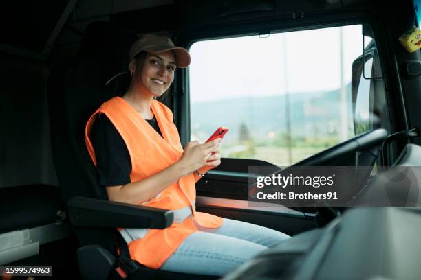 woman truck driver in reflective vest and cap sitting in drivers seat with smart phone - trucker hat stock pictures, royalty-free photos & images