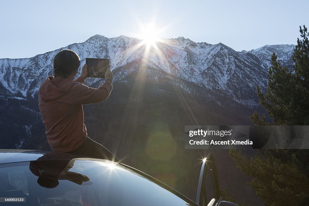 Young man sits on car roof, take pic with tablet