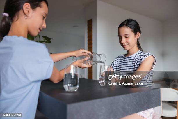 young multiracial girl serves a glass of water to her sister at a kitchen - girl filling water glass stock pictures, royalty-free photos & images