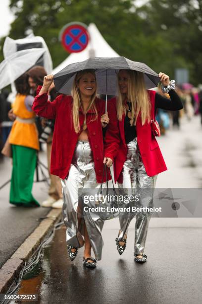 Two guests wear a black umbrella, a red blazer jacket, a black top, silver shiny leather pants, a silver shiny leather handbag, black shiny leather...