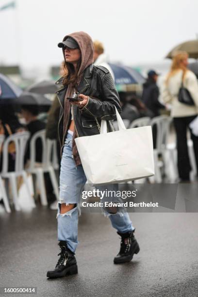 Guest wears a black cap, a dark brown hoodie zipper sweater, a black shiny leather zipper jacket, pale bue ripped knees large pants, a white shopping...