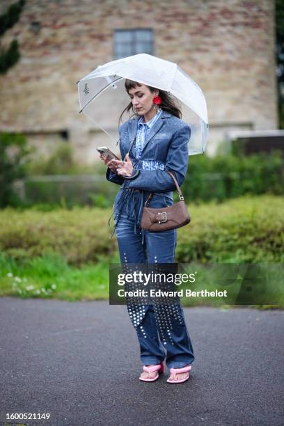 Mari Alexander wears a white transparent umbrella, red large earrings, a white and pale blue striped print pattern shirt, a navy blue denim with...