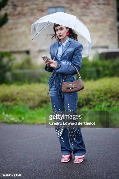 Mari Alexander wears a white transparent umbrella, red large earrings, a white and pale blue striped print pattern shirt, a navy blue denim with...