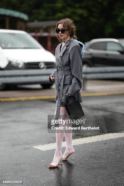 Guest wears black sunglasses, silver earrings, a pale blue feather hat, a gray long blazer jacket with a nailed / studded belt, a black matte leather...