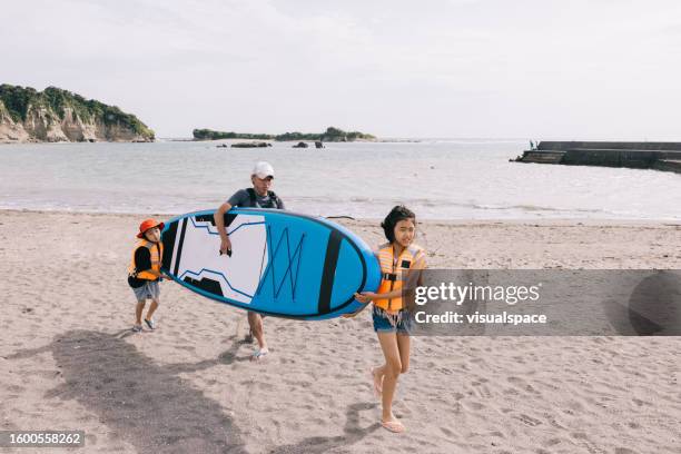 a japanese dad and his children carry a paddleboard to the water - paddleboarding team stock pictures, royalty-free photos & images