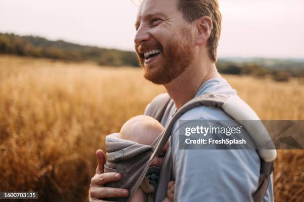 portrait of a caucasian father holding his baby in a chest carrier as he's walking across a field - serbia stock pictures, royalty-free photos & images