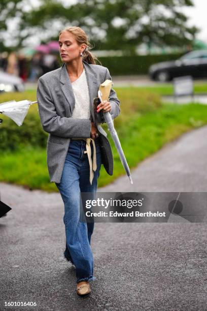 Guest wears silver earrings, a white embroidered sequined asymmetric collar top, a gray blazer jacket, blue denim faded flared pants, silver rings, a...