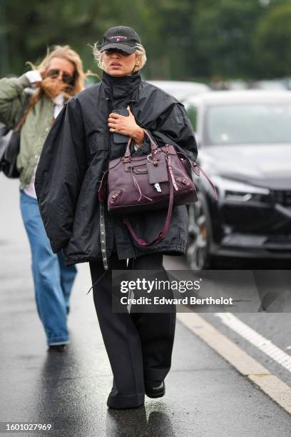 Guest wears a black denim with white and red pattern details cap, a black high neck zipper oversized rain coat, a burgundy matte leather City handbag...