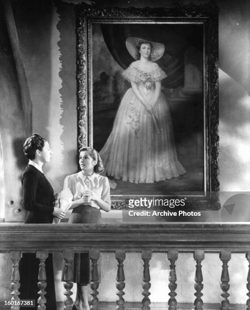 Joan Fontaine and Judith Anderson stand next to a portrait of a woman as they talk in a scene from the film 'Rebecca', 1940.