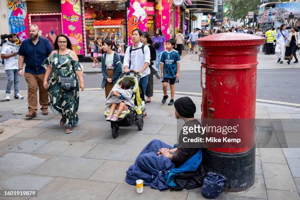 Shoppers and visitors at Leicester Square pass a man sitting underneath a thin sleeping bag on the street on 14th August 2023 in London, United...