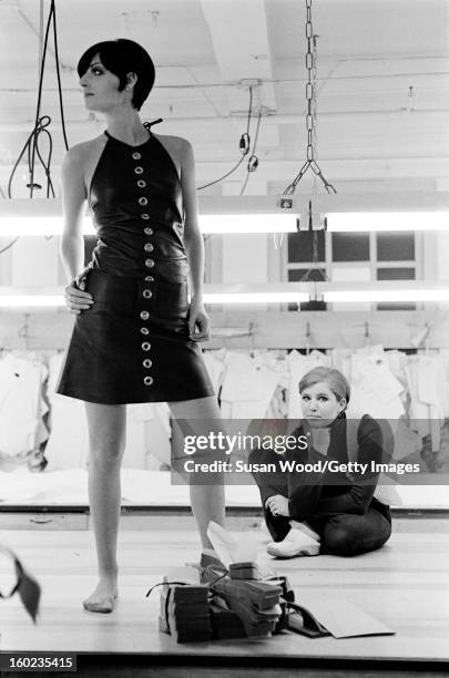 American fashion designer Betsey Johnson sits on the floor of her studio as an unidentified model poses in front of her, New York, New York,...