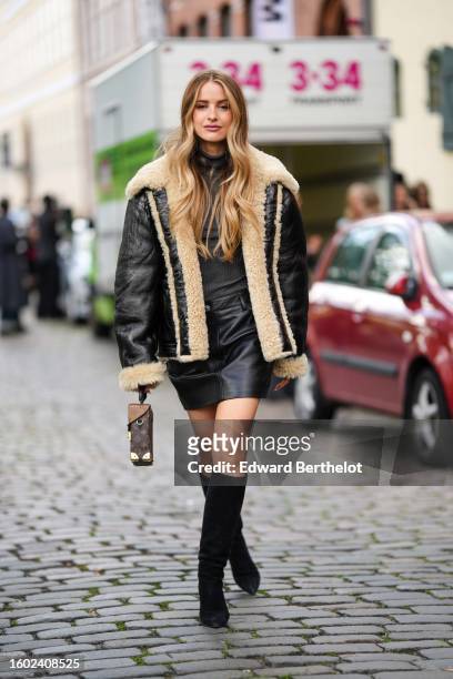 Victoria Magrath wears a dark gray ribbed wool turtleneck pullover, a black shiny leather with beige sheep fur details oversized aviator coat, a...