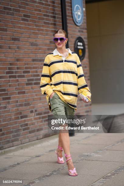 Marianne Theodorsen wears purple large aviator sunglasses, a yellow with black large striped print pattern oversized polo shirt, khaki cargo knees...