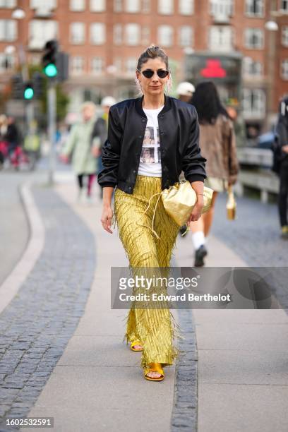 Guest wears black sunglasses, a white t-shirt with pictures of Kim Kardashian print pattern t-shirt, a black satin silk bomber jacket, gold...