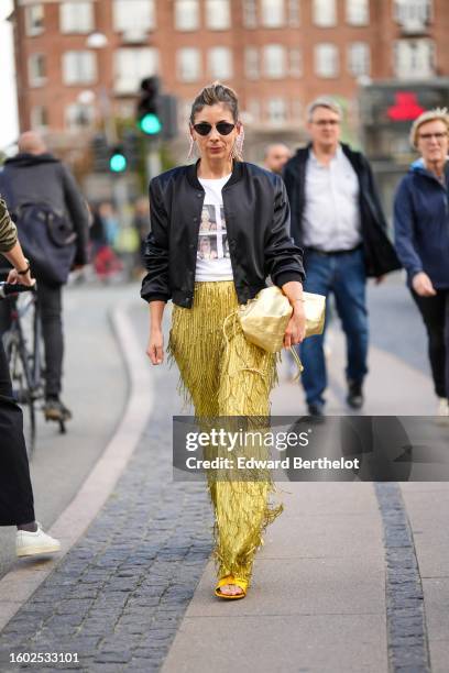 Guest wears black sunglasses, a white t-shirt with pictures of Kim Kardashian print pattern t-shirt, a black satin silk bomber jacket, gold...