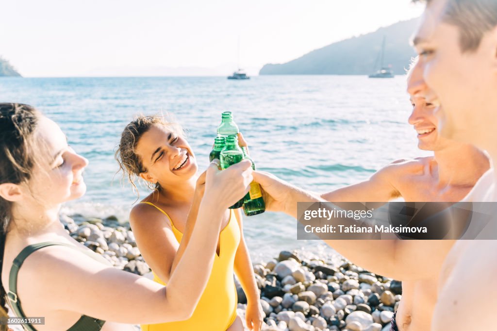 Group of mid adult friends has a fun with bottles of drinks on the beach. Sea and sunset on the background.