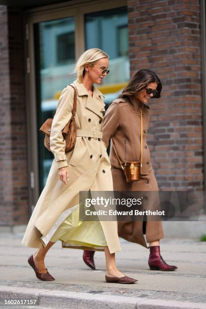 Guest wears black small circle sunglasses, a beige long belted trench coat, a brown suede large shoulder bag, a pale yellow silk long dress, dark...