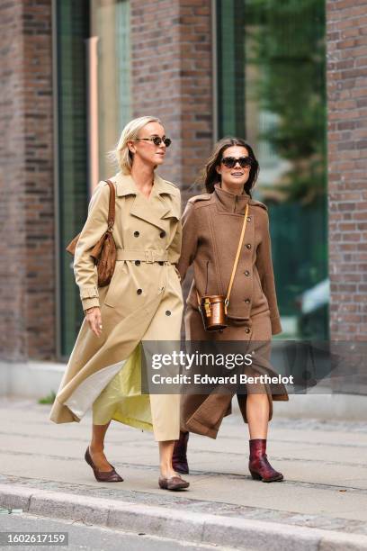 Guest wears black small circle sunglasses, a beige long belted trench coat, a brown suede large shoulder bag, a pale yellow silk long dress, dark...