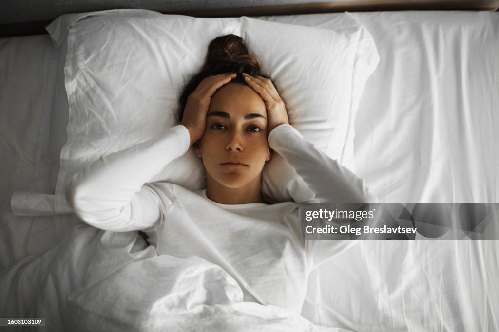 Sad woman lying down in bed with migraine or headache. Health problem, fair, insomnia or panic attack