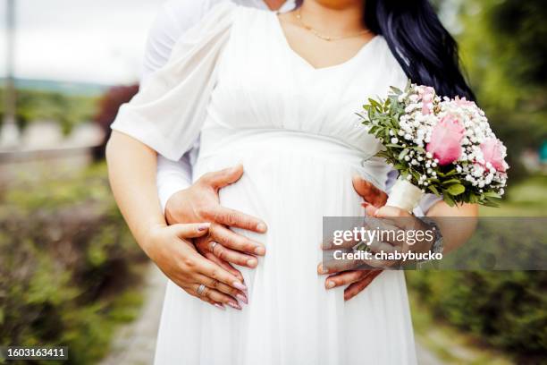 schwangere braut hält blumen, während ehemann ihren bauch hält - white dress stock-fotos und bilder